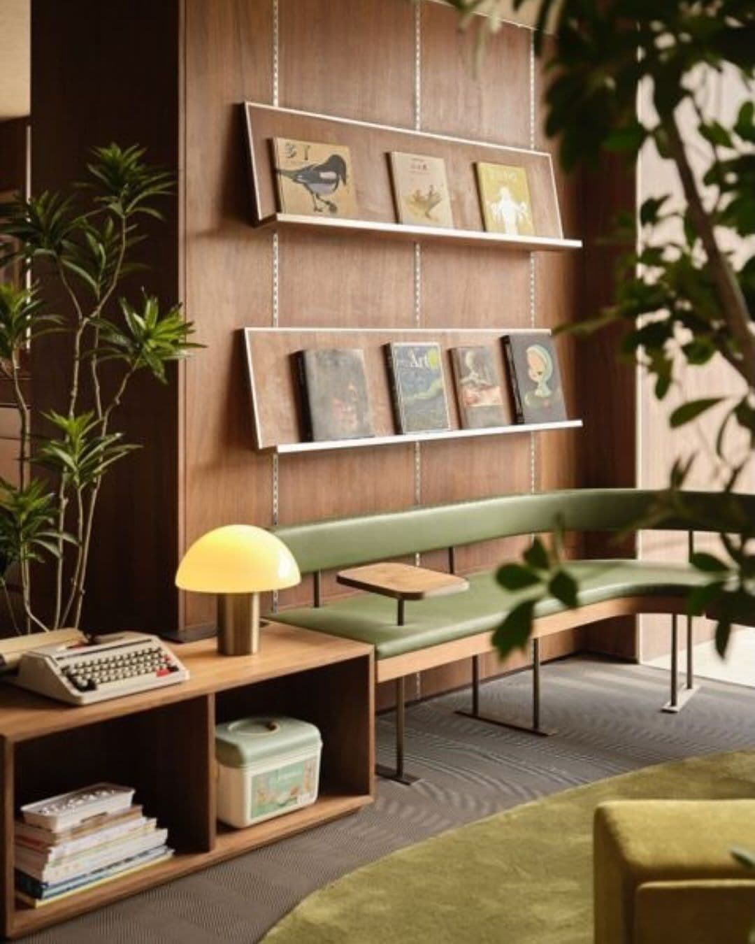 Rincón de lectura con una banca verde, una lámpara de mesa y revisteros de madera en la pared