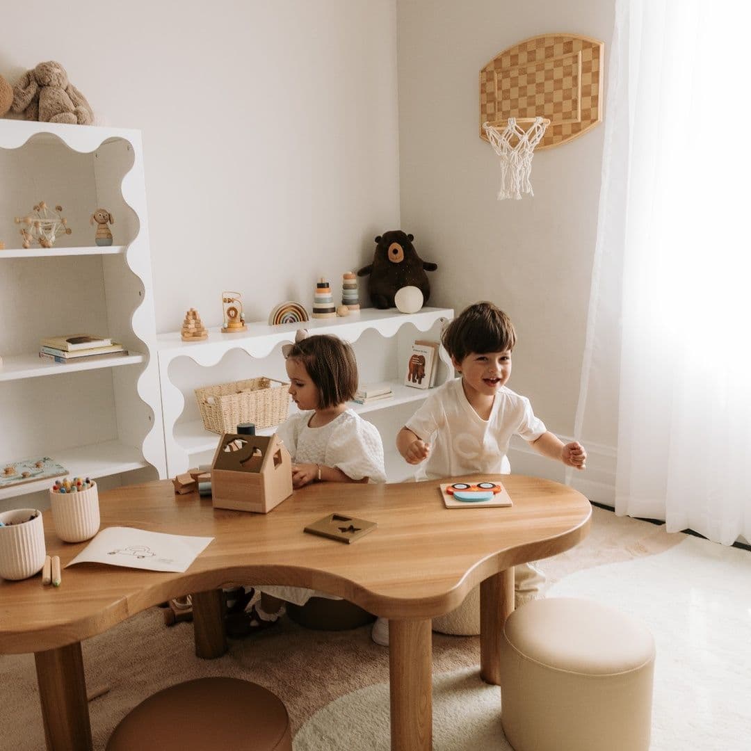 Dos niños jugando con juguetes de madera en una mesa curva, en una sala de juegos con estantes ondulados y un aro de básquetbol de mimbre