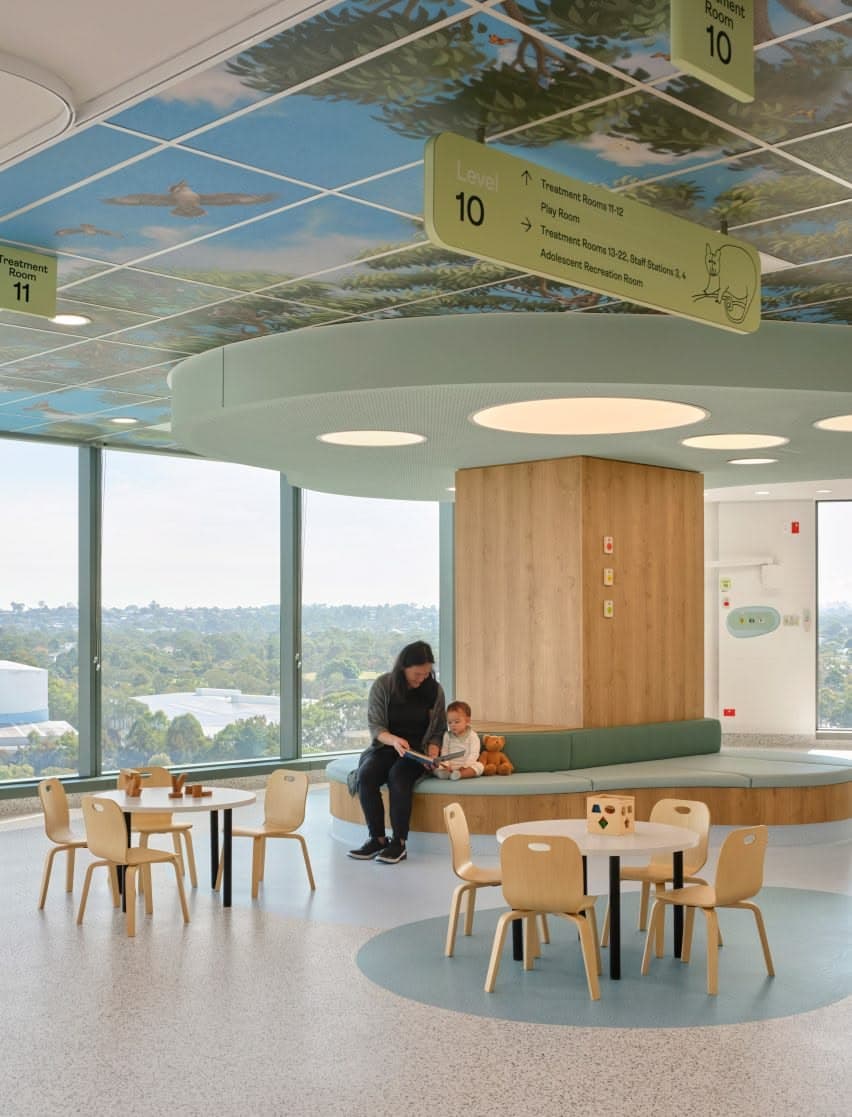 Sala de espera pediátrica con un cielo pintado de árboles, banca curva y mesas infantiles
